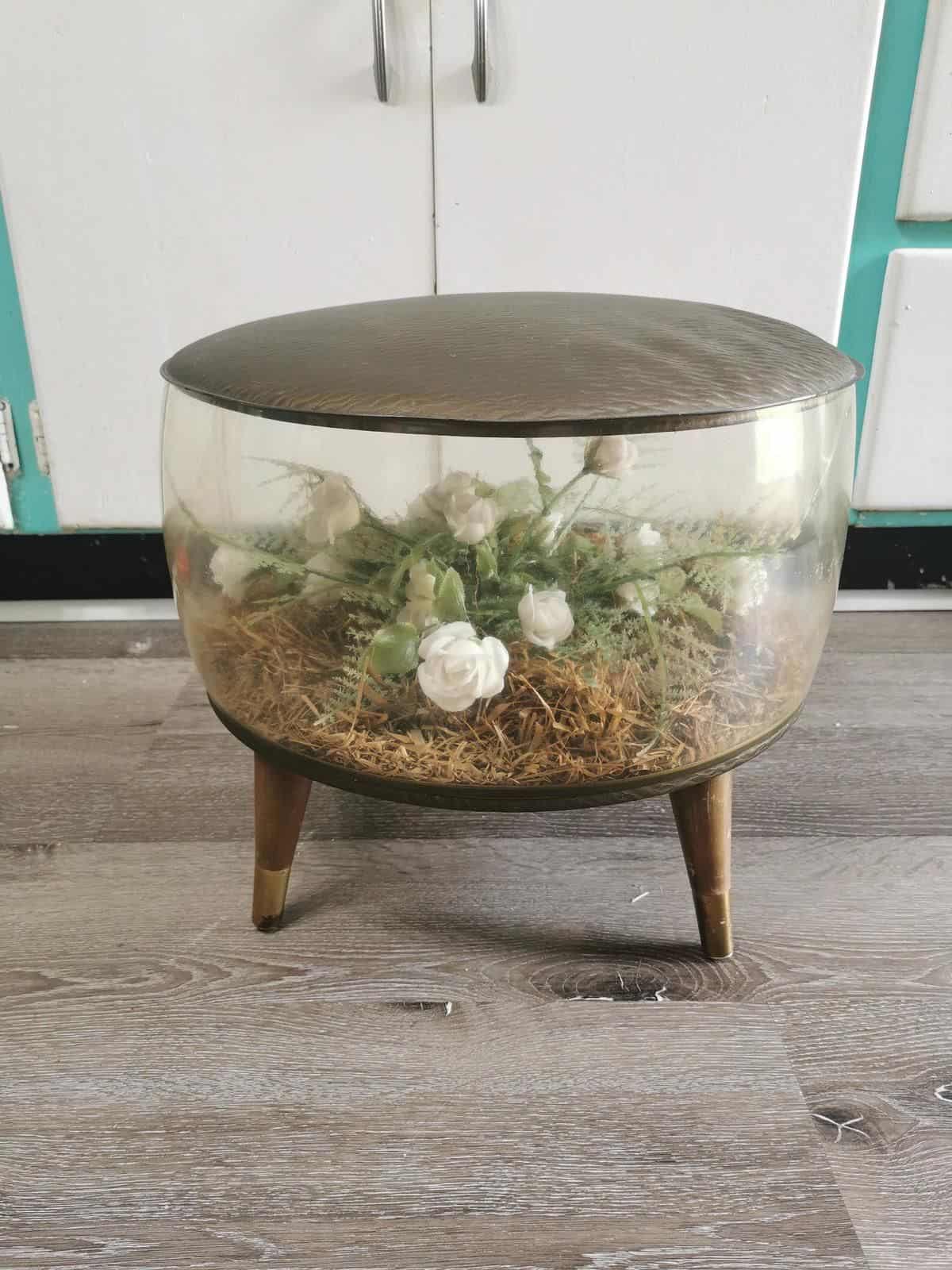 Transparent vintage circular ottoman with brown top and faux plants and flowers inside